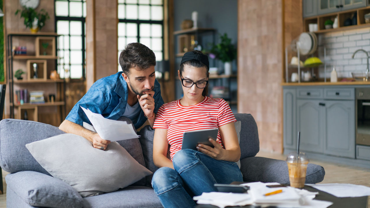 Couple calculating bills at home using laptop and calculator. Young couple working on computer while calculating finances sitting on couch. Young  man with  wife at home analyzing their finance with documents.