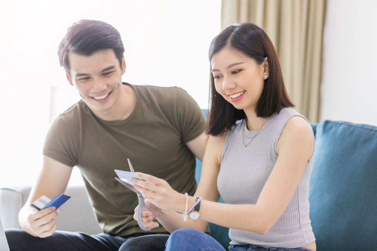 A young woman with scissors, cuts a credit card while smiling. Her husband holds more cards and smiles. They are both sitting in their living room.