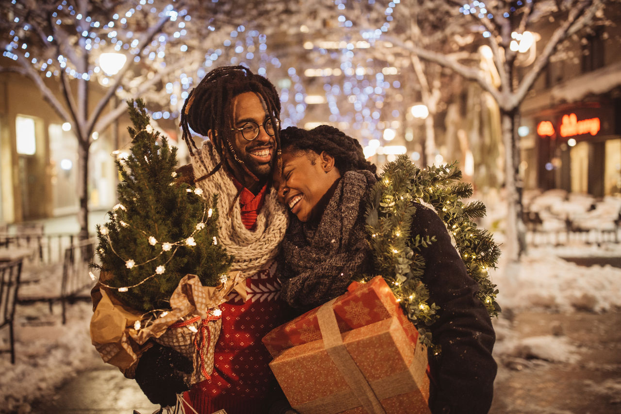 Young couple preparing for Christmas holiday. They walking on street and holding gift boxes and Christmas tree