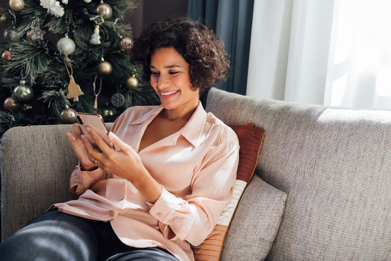 Smiling female sitting on couch next to the Christmas tree and typing text message on her smartphone