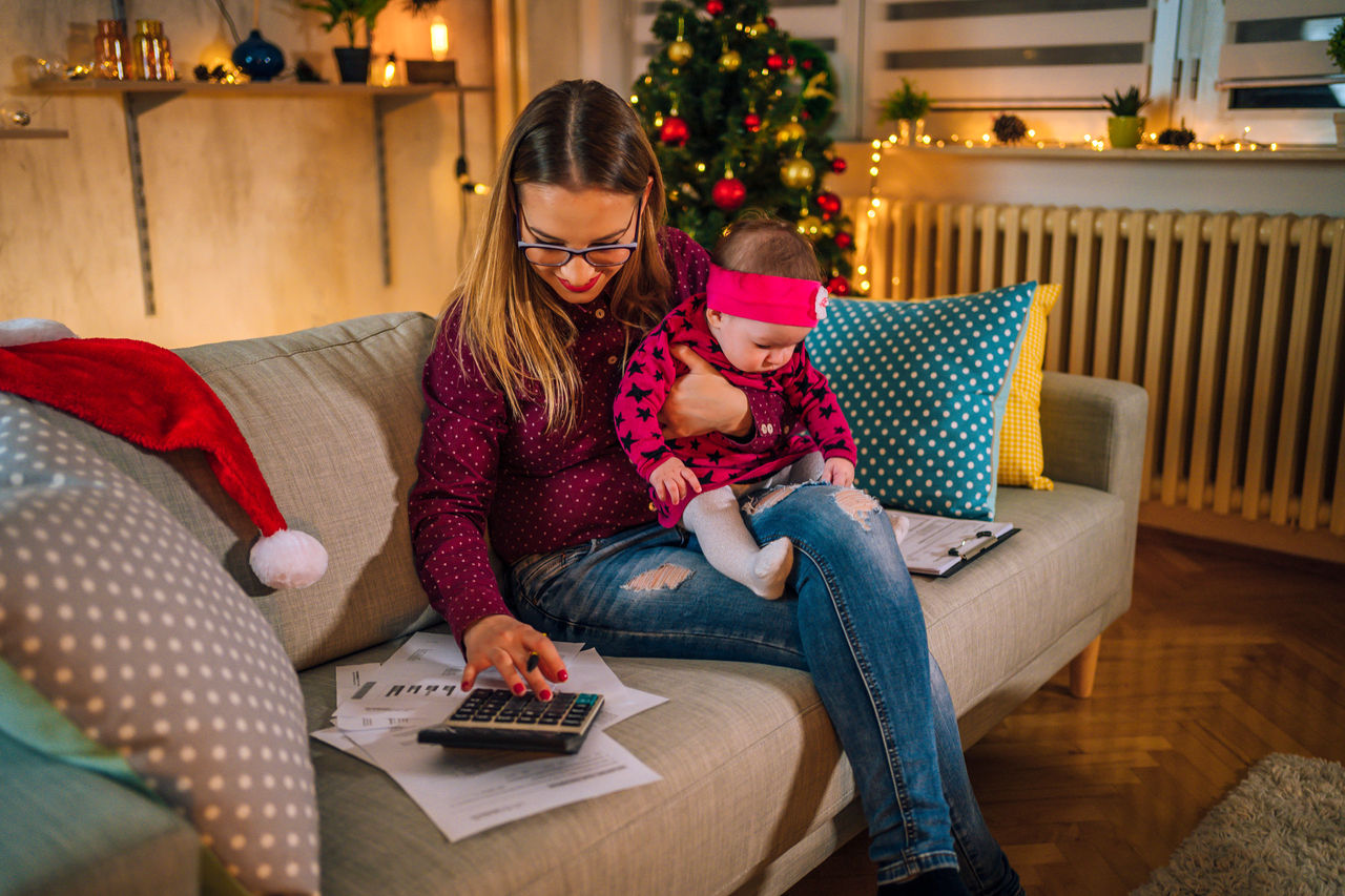 Young cheerful mother with a baby in her arms managing finances in the living room during Christmas holidays