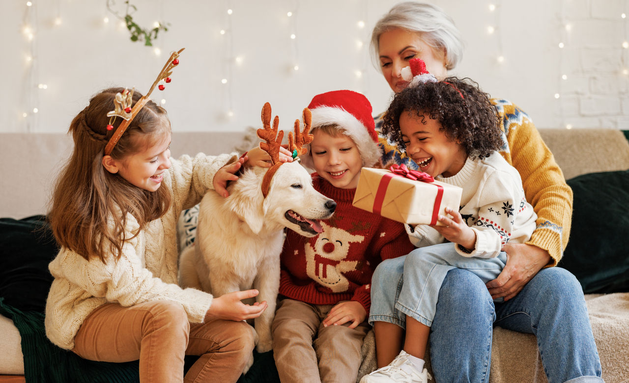 Happy multiethnic family with dog on Christmas day, little kids siblings of different races spend winter holidays at grandma house. Loving senior grandmother receiving New Year gift from grandchildren