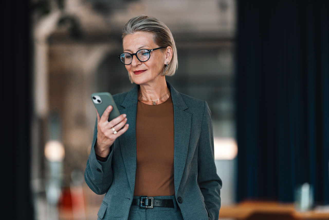 Close up shot of a beautiful senior woman reading a message on her mobile phone. She is standing in the office, looking down at the screen and smiling.
