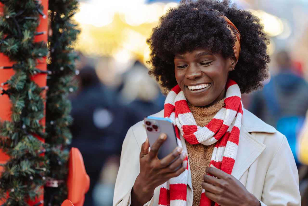 A cheerful woman with curly hair and a striped scarf enjoys the festive atmosphere outdoors, holding a smartphone in a holiday setting.
