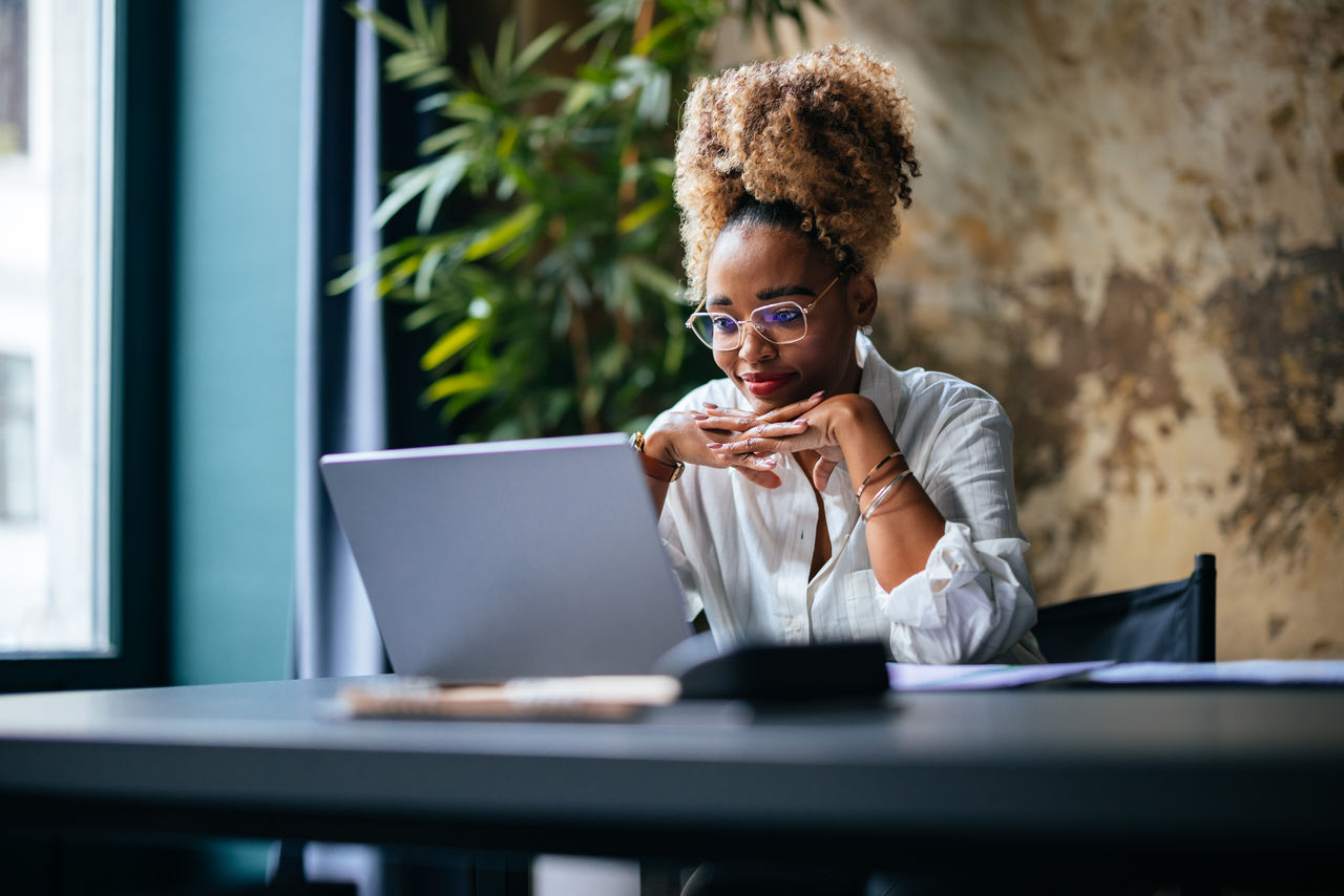 Happy African-American woman reading business report on her laptop while sitting at restaurant desk.