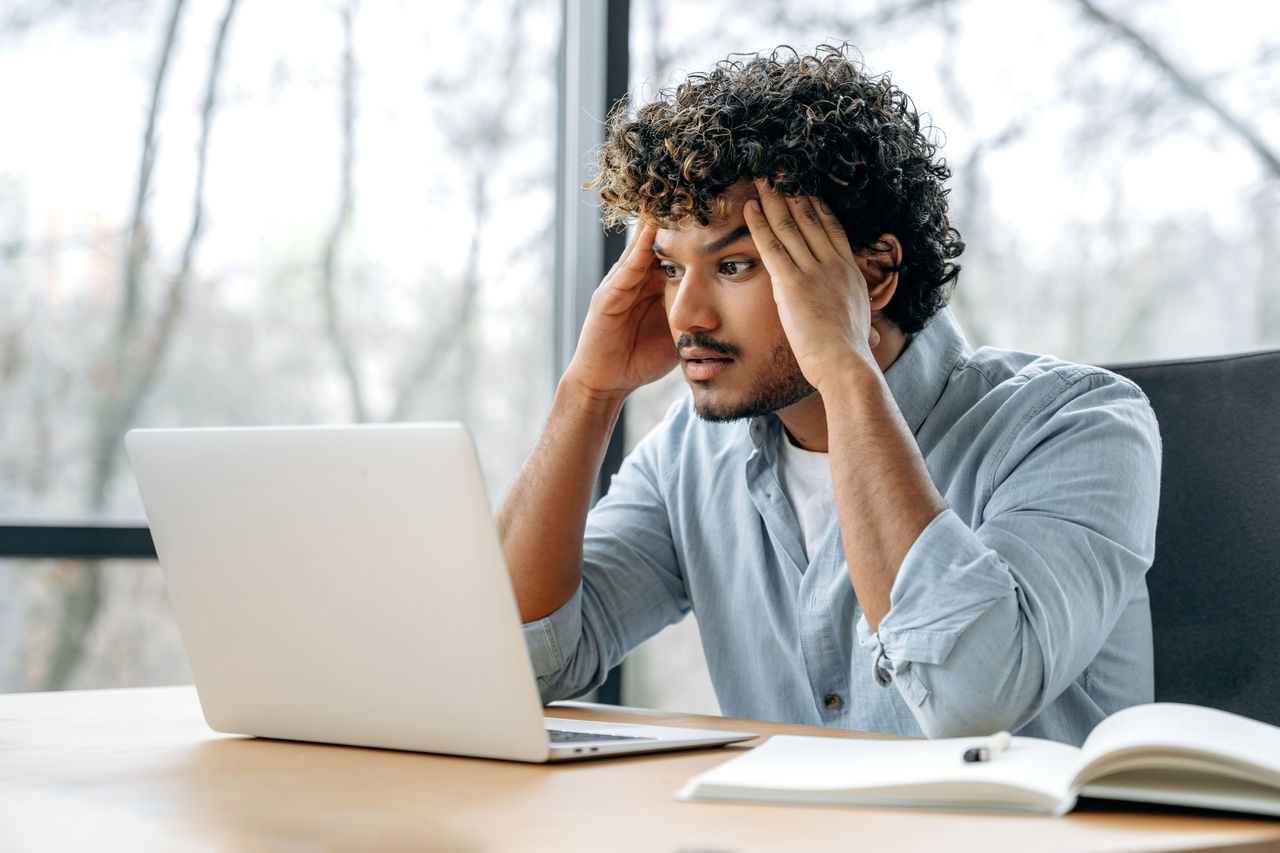 Tired exhausted overworked indian office worker, creative manager, sitting at workplace holding his hands to his head, feeling stressed and tired from long work in laptop, needs a break, rest