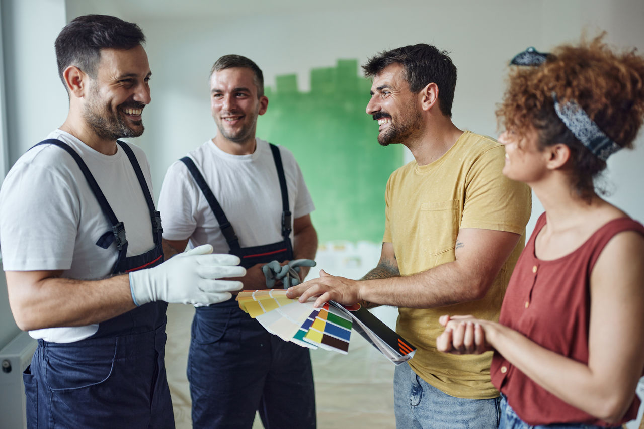 Young happy couple talking to house painters while choosing colors during home renovation process.