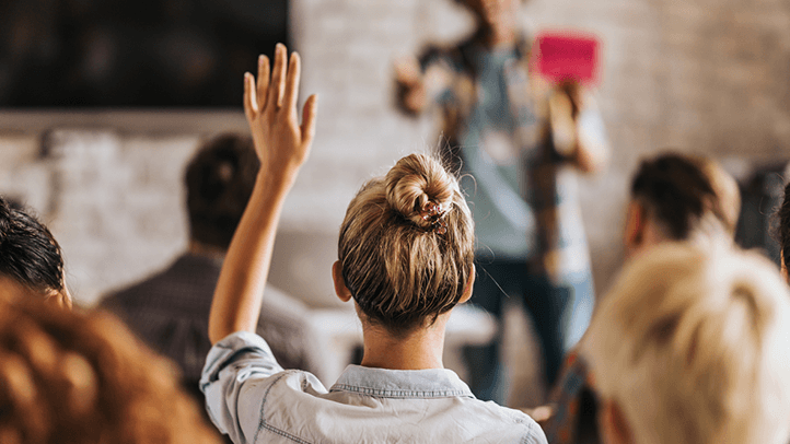 Woman sitting in crowd at a speaking event with her hand raised