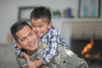 A military dad and his son are hugging in their living room. The son is smiling happily at the camera.