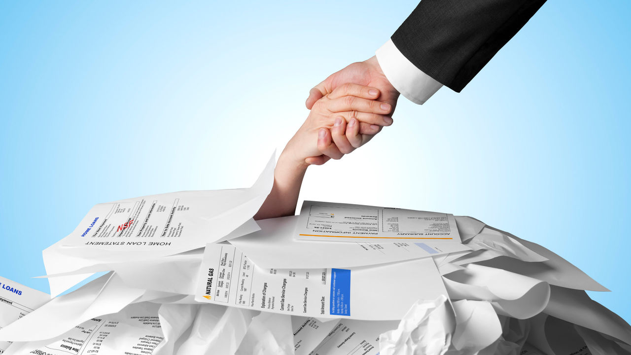 a hand reaching out from under a pile of paper bills and grasping onto a suited hand 