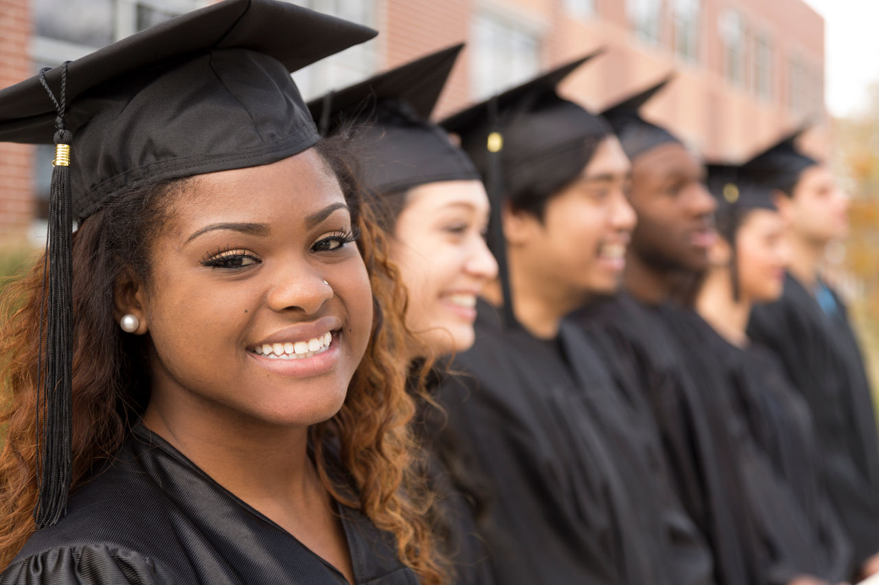Six multi-ethnic friend graduates excitedly wait for their name to be called during graduation ceremony. African descent girl looks back at camera. School building background.