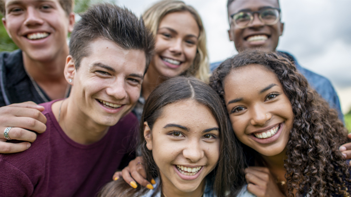 A group of eight multi-ethnic teenagers, 17 and 18 years old, carrying book bags, standing together outside a school building. They are high school seniors or university freshmen.