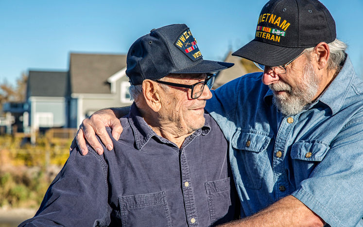 Two generations of real person senior adult men USA military veterans. On the left, a 93 year old United States Army World War II and Korean Conflict US Air Force military veteran is looking at his son-in-law on the right. He's a 65 year old US Navy sailor Vietnam War military veteran. The younger man is teasing his father-in-law - trying to distract him by telling jokes during the photo shoot. They are both wearing their own era-specific generic souvenir shop military veteran commemorative baseball style caps.