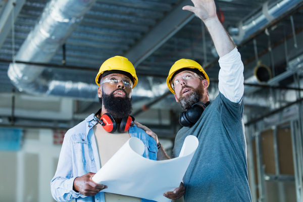 Two multi-ethnic construction workers wearing hard hats and safety glasses at an indoor construction site. They are both mature men in their 40s with thick beards. The African-American man is holding a set of floor plans. The structure being built is an office building.