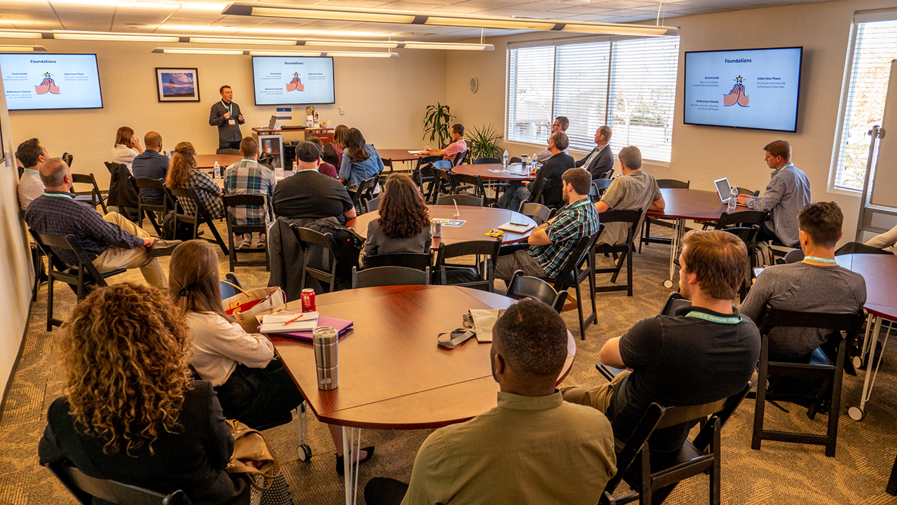 large room filled with many people sitting at tables while a speaker gives a presentation at the front of the room