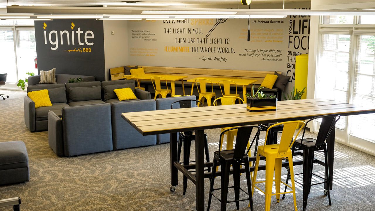 open coworking area with grey couches, yellow table, and a bench along the wall, and various famous quotes on the walls, and a long industrial table with tall black and yellow chairs