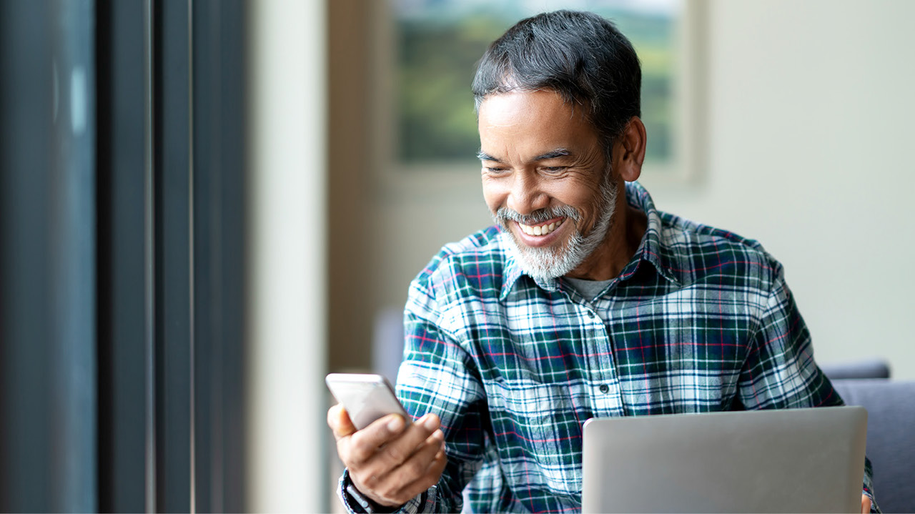 Photo of older man, smiling while using computer and phone
