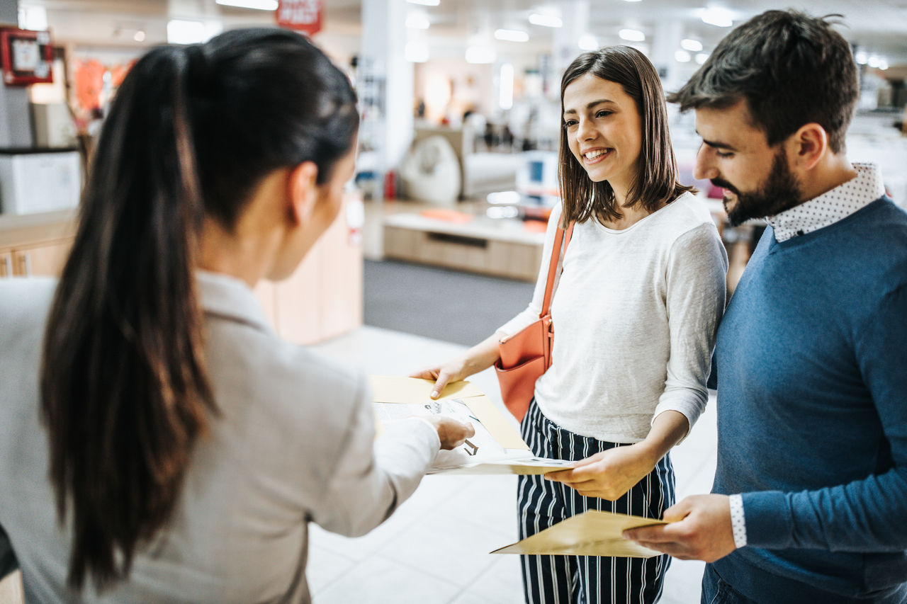 Young couple and saleswoman looking for choices in catalog at furniture store. Focus is on happy woman.