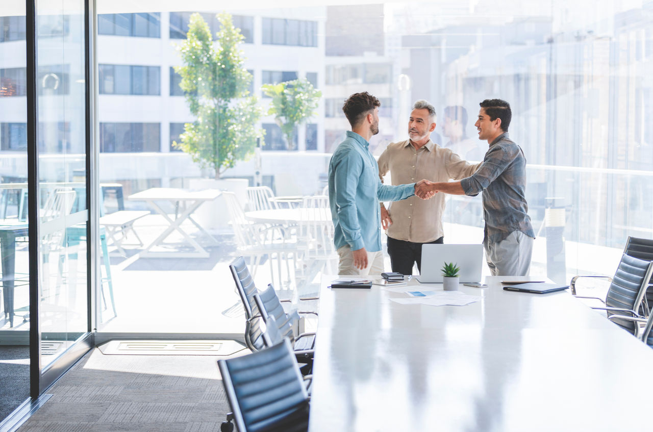 Three businessmen shaking hands.  They are casually dressed and one man is mature with grey hair. They are discussing something at a conference table that has some technology and paperwork on it.