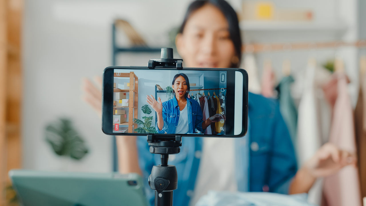 1320976219 Young Asia lady fashion designer using mobile phone receiving purchase order and showing clothes recording video live streaming online at shop. Small business owner, online market delivery concept.