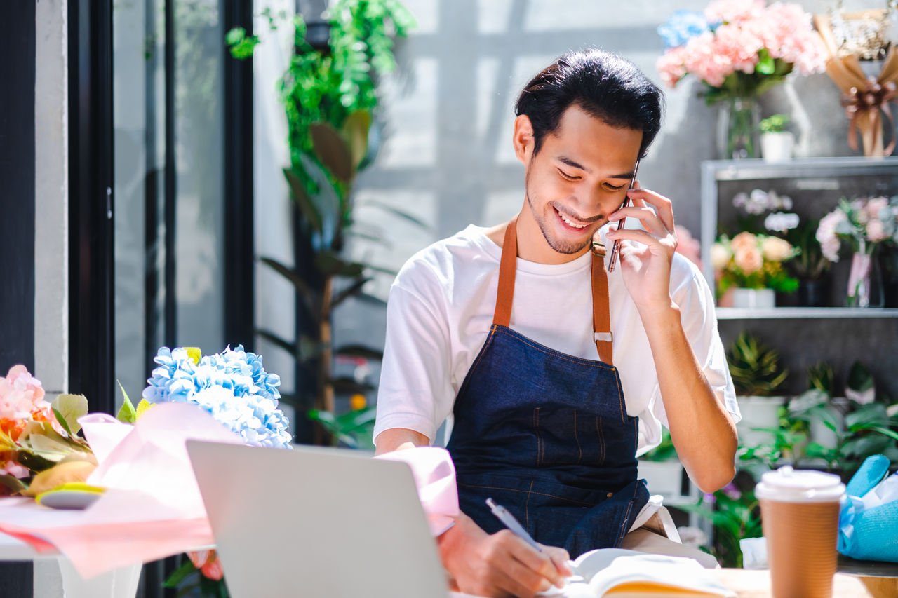 1422731163 Asian man influencer or SME owner people smile work on home video camera selfie shoot.Smiling female florist talking on mobile phone. Young woman working at shop with Open sign.