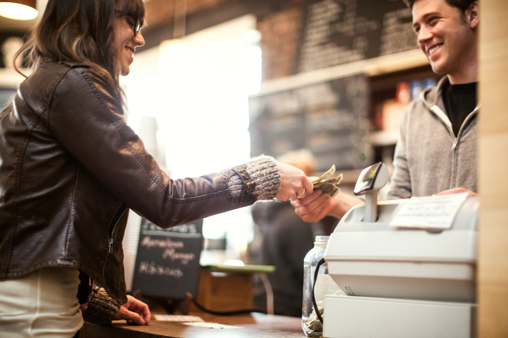 A woman is paying at a coffee shop.  The cashier is a young man.  They are smiling at each other.